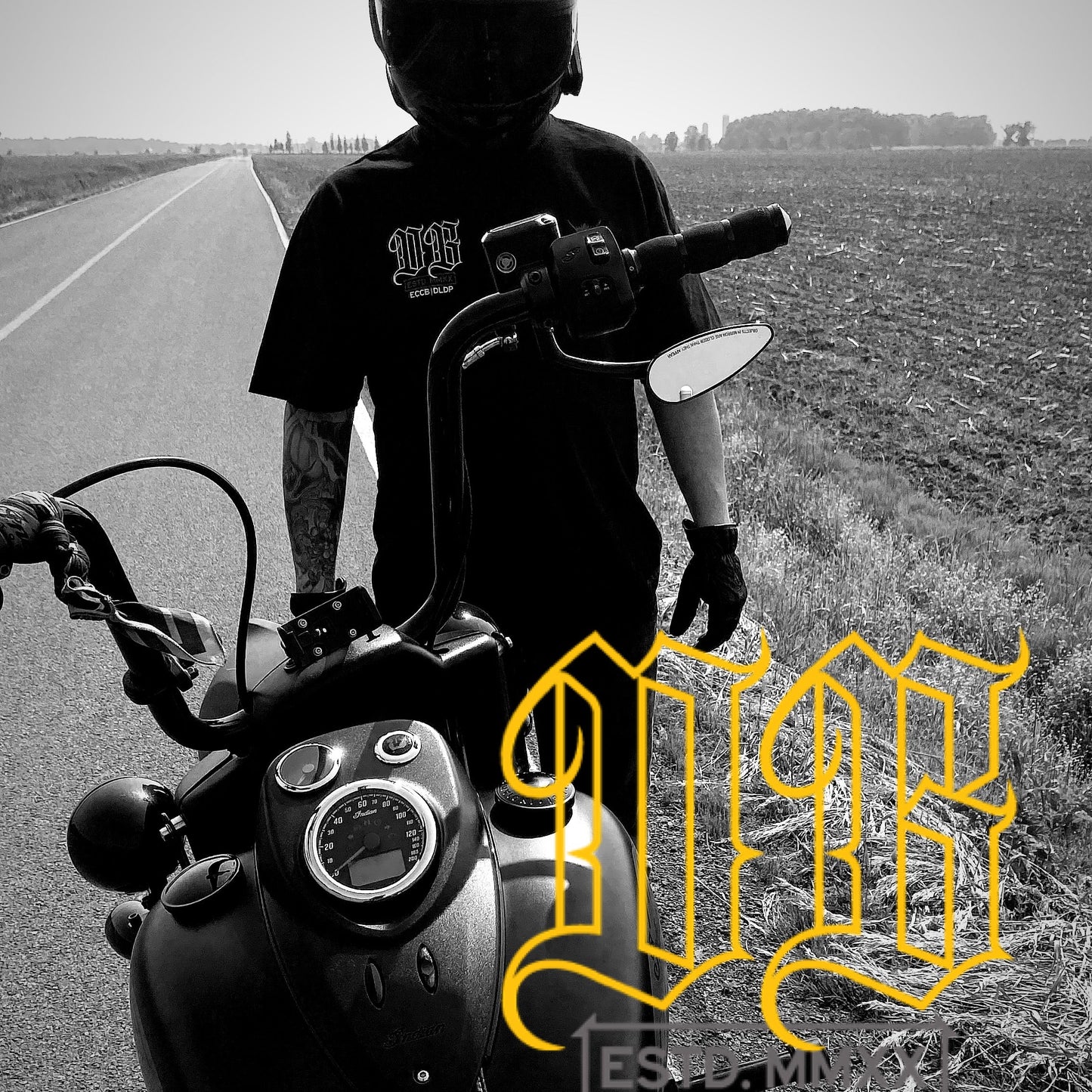 Person standing by motorcycle with a helmet, standing on a road with fields in the background.